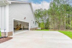 A two-car garage with an open door in a gray house, adjacent to a large paved driveway and a grassy area with tall trees in the background.