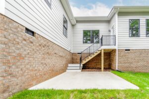 Backyard view of a house with brick and white siding, showing a small staircase leading to a narrow elevated balcony and a concrete patio area surrounded by grass.