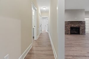A hallway with light beige walls and wooden flooring, leading to white doors. A stone fireplace is visible in the adjacent room on the right.