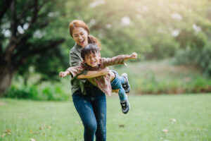 A woman twirls a smiling child in the air while standing on a grassy field with trees in the background.