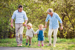 Two elderly adults walk with two young children outdoors on a clear day, surrounded by greenery. The adults hold the children's hands as they walk on a path.