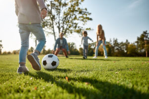 A child kicks a soccer ball across the grassy field of Poplar Village, with three people enjoying the sunny day in the background.