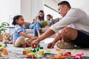A family is in a living room. A man and a young girl are playing with toys on the floor, while a woman reads to another girl on the couch.
