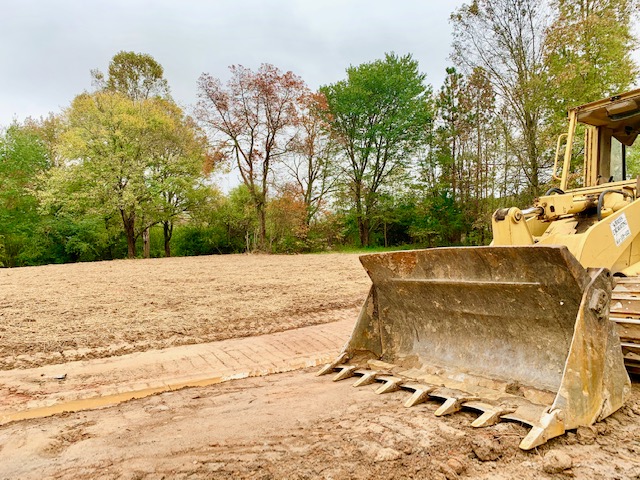 A large bulldozer with a metal blade parked on dirt ground in front of a cleared area, with various trees and greenery in the background.