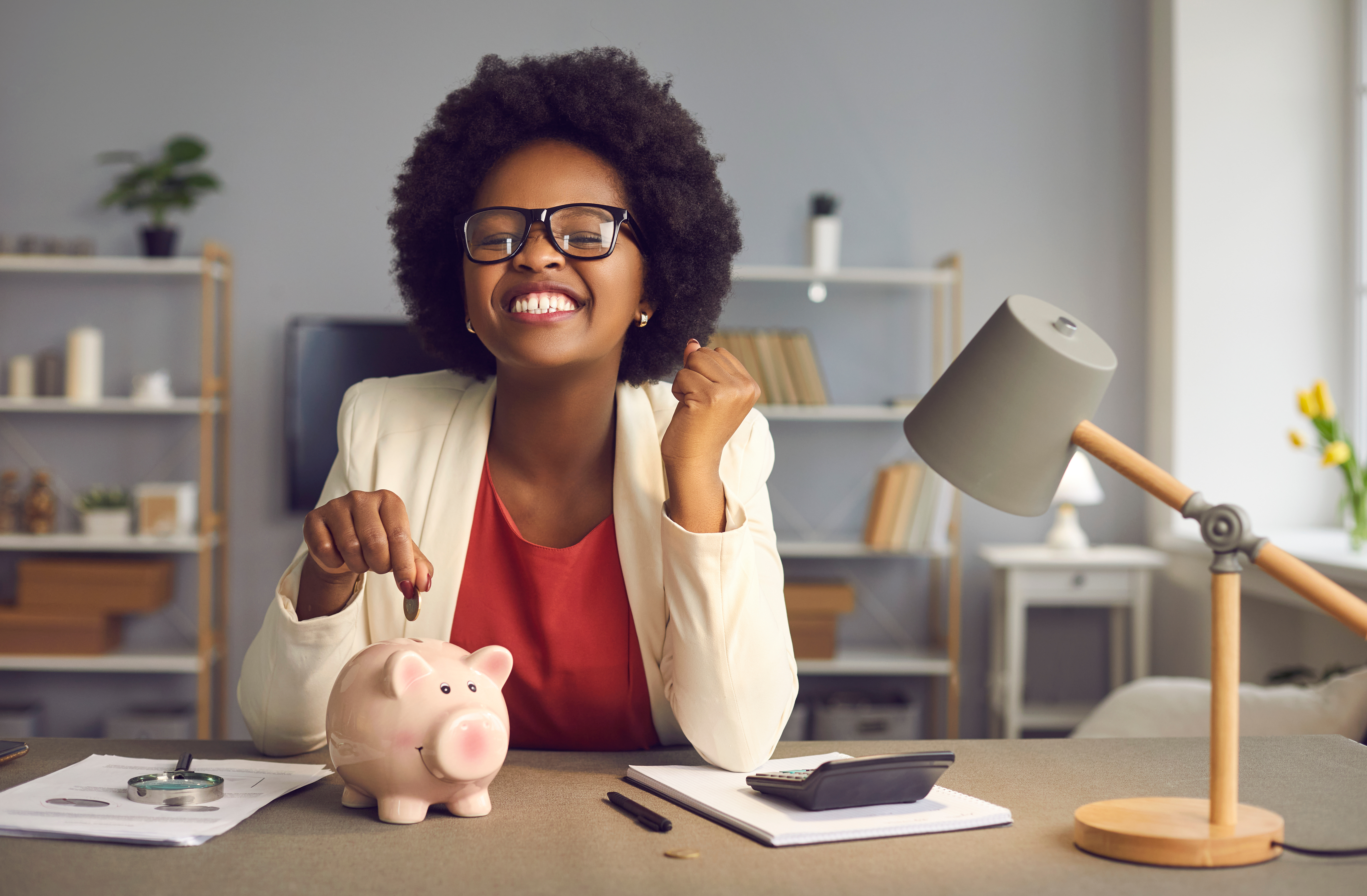A woman with curly hair and glasses, wearing a white blazer, smiles while placing a coin into a pink piggy bank on a desk with papers, a calculator, and a lamp.
