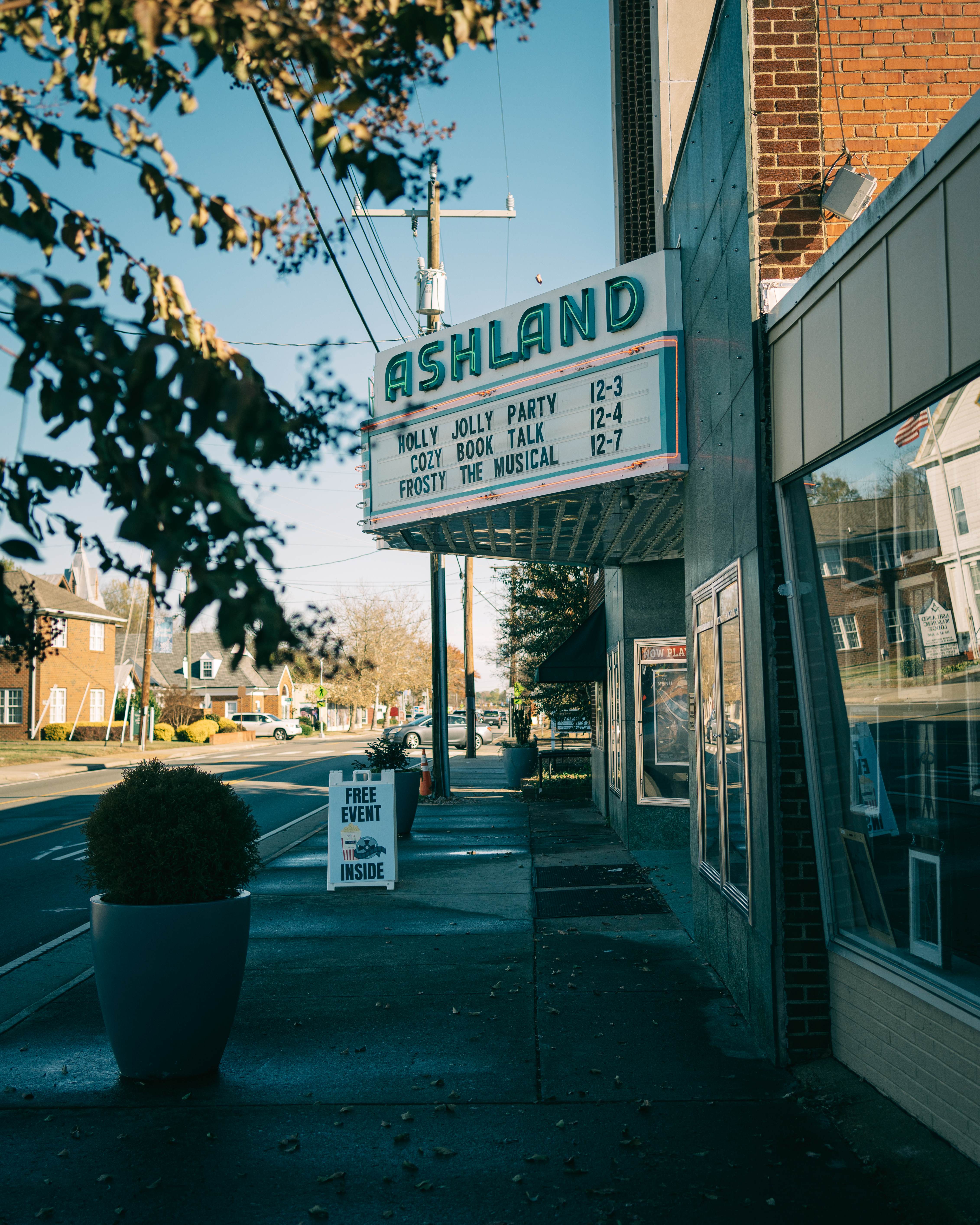 Street view of a theater with a marquee displaying event titles and times, and a sign below advertising a free event inside. Trees and buildings are visible in the background.