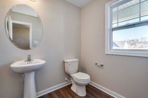 Minimalist bathroom featuring a round mirror and pedestal sink, accompanied by a sleek toilet. Sunlight streams through the window on the right, illuminating the warm wood flooring. It feels like a serene station of relaxation amidst Carter's refined design elements.