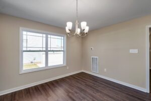 The empty dining room, reminiscent of a classic dispatch station, features a wooden floor, beige walls, and a large window. A modern chandelier hangs overhead, casting elegant light throughout the space.