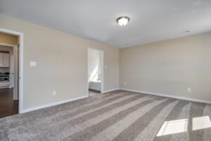 Empty room with beige walls, carpeted floor, and ceiling light. Doorways seamlessly connect to a Carter kitchen and a sunlit hallway resembling a tranquil dispatch station.