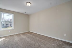 The empty room, once a bustling dispatch station, now offers a serene setting with its beige walls and carpeted floor. Natural light streams in through the window, while the ceiling light fixture stands ready to illuminate any moment's transition.