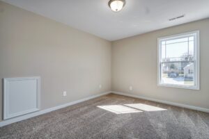 An empty room with beige walls and a carpeted floor sits quietly, reminiscent of an old station waiting for life. A single window welcomes natural light, while the ceiling light fixture stands by like a dispatch ready to illuminate the space.