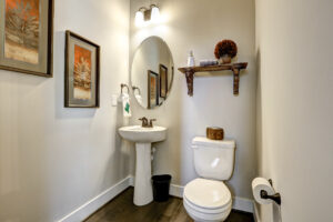 The bathroom features a Westwood pedestal sink, round mirror, toilet, and shelf. Framed art and wooden accents complement the cream walls, creating an elegant yet cozy space reminiscent of a classic Dispatch Station.