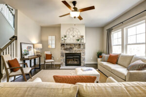 Cozy living room with beige sofas and a stone fireplace, reminiscent of a Westwood retreat. A ceiling fan spins leisurely under the bright natural light, offering both comfort and charm akin to an inviting dispatch station ambiance.