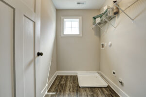 The Westwood laundry room features a small window, wire shelves, and plumbing hookups on a wooden floor—your personal dispatch station for laundry day efficiency.