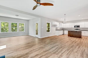Discover the open-concept kitchen and living area in Lot 37, featuring wooden floors, sleek white cabinets, and a ceiling fan. Nestled in Brickshire, this inviting space offers calming views of lush green trees.