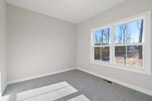 The empty room at Lot 19 features a light gray carpet, beige walls, and a large window offering a view of trees and the road. It's a perfect spot to enjoy the serene atmosphere of Autumn Hill.