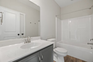 The minimalist bathroom in Lot 19 at Autumn Hill features a white bathtub, shower, sink, and a sleek gray vanity. Warm wood flooring enhances the serene ambiance.