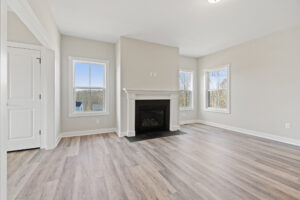 The empty room on Lot 19 features light gray walls, wood flooring, and a white fireplace. Three large windows flood the space with natural light, creating a serene atmosphere in the heart of Autumn Hill.