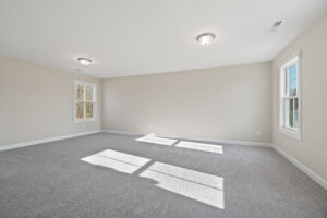 The empty room at Lot 19 features beige walls, a gray carpet, and two windows that welcome natural light. Nestled in the serene setting of Autumn Hill, the ceiling lights glow warmly, creating a cozy atmosphere.