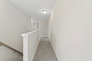 A modern hallway in Autumn Hill features beige walls, a carpeted floor, and a white railing. It leads to a door with a ceiling light fixture, offering a glimpse into the elegant design of Lot 19.