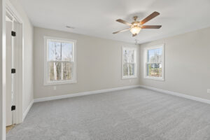 The bright, empty room in Autumn Hill features beige walls, gray carpet, a ceiling fan, and three windows that offer serene views of the trees outside. It's a perfect spot on Lot 19 to enjoy the tranquility of nature right at your doorstep.