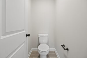 The white toilet sits in a small, light gray bathroom on a wood-patterned floor with simple fixtures, capturing the cozy charm of Lot 19 at Autumn Hill.