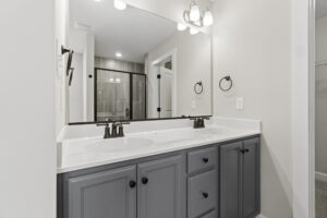 The Autumn Hill modern bathroom features a dual sink vanity, a large mirror, and a glass shower enclosure with a sleek gray and white color scheme. Located at Lot 19, this space exudes elegance and style.