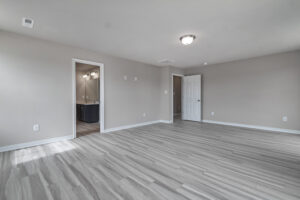 A spacious empty room with light gray walls and wood flooring, featuring a white door, a ceiling light fixture, and a doorway leading to a bathroom with double sinks.