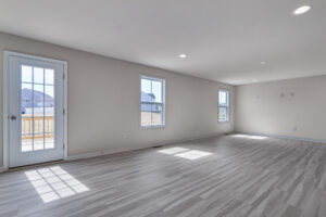 An empty, well-lit room with light wood flooring, two windows, and a glass door leading outside. Recessed lighting is installed in the ceiling.