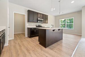 A modern kitchen with wooden flooring, dark cabinets, an island, stainless steel appliances, and pendant lighting. A large window provides natural light from the outside.