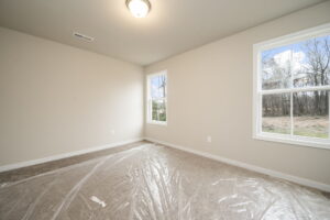 Empty room with beige walls, a light fixture on the ceiling, two windows showing an outdoor wooded area, and a floor covered in protective plastic.