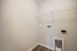 Empty laundry room with a wire shelf, utility connections, and beige walls. Wood flooring and baseboard trim are visible.
