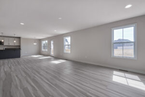 A spacious, empty living area with light-colored wooden floors, several windows, and a view of a modern kitchen with hanging lights in the background.