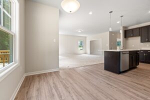 An empty, open-concept living space with hardwood floors, beige walls, and a modern kitchen featuring dark cabinets and an island. Large windows provide natural light.