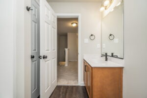A bathroom with a single vanity, mirror, and light fixture. There are double doors to the left and an open doorway leading to a hallway. The floor is a mix of dark wood and light tile.