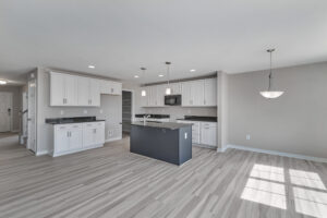 A modern kitchen with white cabinets, a gray island, stainless steel appliances, and light wood flooring. A pendant light hangs over the dining area, with large windows letting in natural light.