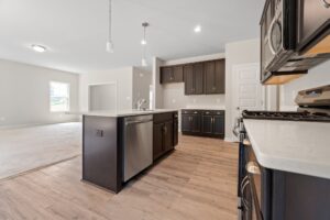 Modern kitchen with dark wood cabinets, stainless steel appliances, and an island with a sink. Open layout extends to a carpeted living area in the background. Hardwood floors throughout.