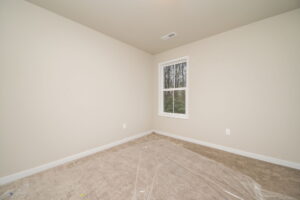 A small, empty room with beige walls and a window overlooking trees. The carpeted floor is covered with a clear plastic sheet.
