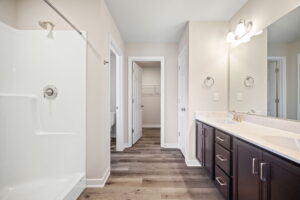 A bathroom with a large mirror, double sink vanity, shower with glass doors, and a closet in the background. The floor is wooden, and the walls are painted a light color.