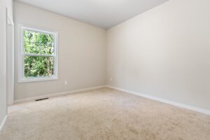 A spacious empty room with beige walls, a carpeted floor, a single window showing greenery outside, and a white baseboard.