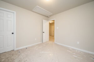An empty room with beige walls, a light-colored carpet covered in plastic, an open door leading to a hallway, and a closed white door. A ceiling hatch is visible.