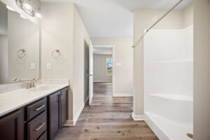 A bathroom interior with light colored walls features a vanity with two sinks, a wall mirror, and a shower area on the right. The floor is hardwood, and a doorway leads to another room.