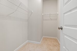 A small walk-in closet with beige carpet, white walls, two white wire shelves, and a white door with a round knob.