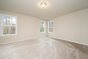 An empty room with beige walls, carpeted floor, and two windows. A ceiling light is on, and some plastic sheeting is spread on the floor.