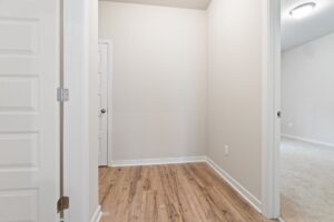 A small hallway with light-colored walls, wooden flooring, a white door on the left, and an open room with carpeted flooring on the right. A ceiling light is visible in the hallway.