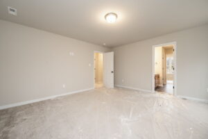 A bright, empty room with plastic covering the light carpet, beige walls, a ceiling light fixture, three doorways, and one of the doors leading to a bathroom.
