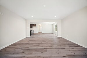 Spacious, empty living room with light gray walls and dark wood flooring, leading to an open kitchen with white cabinetry and stainless steel appliances in the background. Ceiling lights illuminate the space.