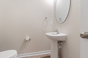 A small bathroom features a toilet, a pedestal sink with a round mirror above it, and a toilet paper holder mounted on the wall, all against a light beige wall.