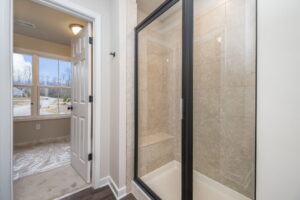 A bathroom with a glass-enclosed shower, beige tiles, and an adjacent open door leading to a carpeted room with a window showing an exterior view.