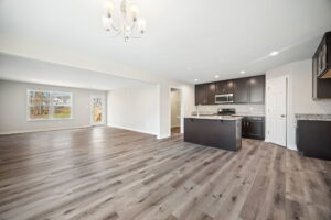 An open-concept living area with hardwood floors, featuring a modern kitchen with dark cabinetry and stainless steel appliances, adjacent to a spacious living room and dining area under a chandelier.